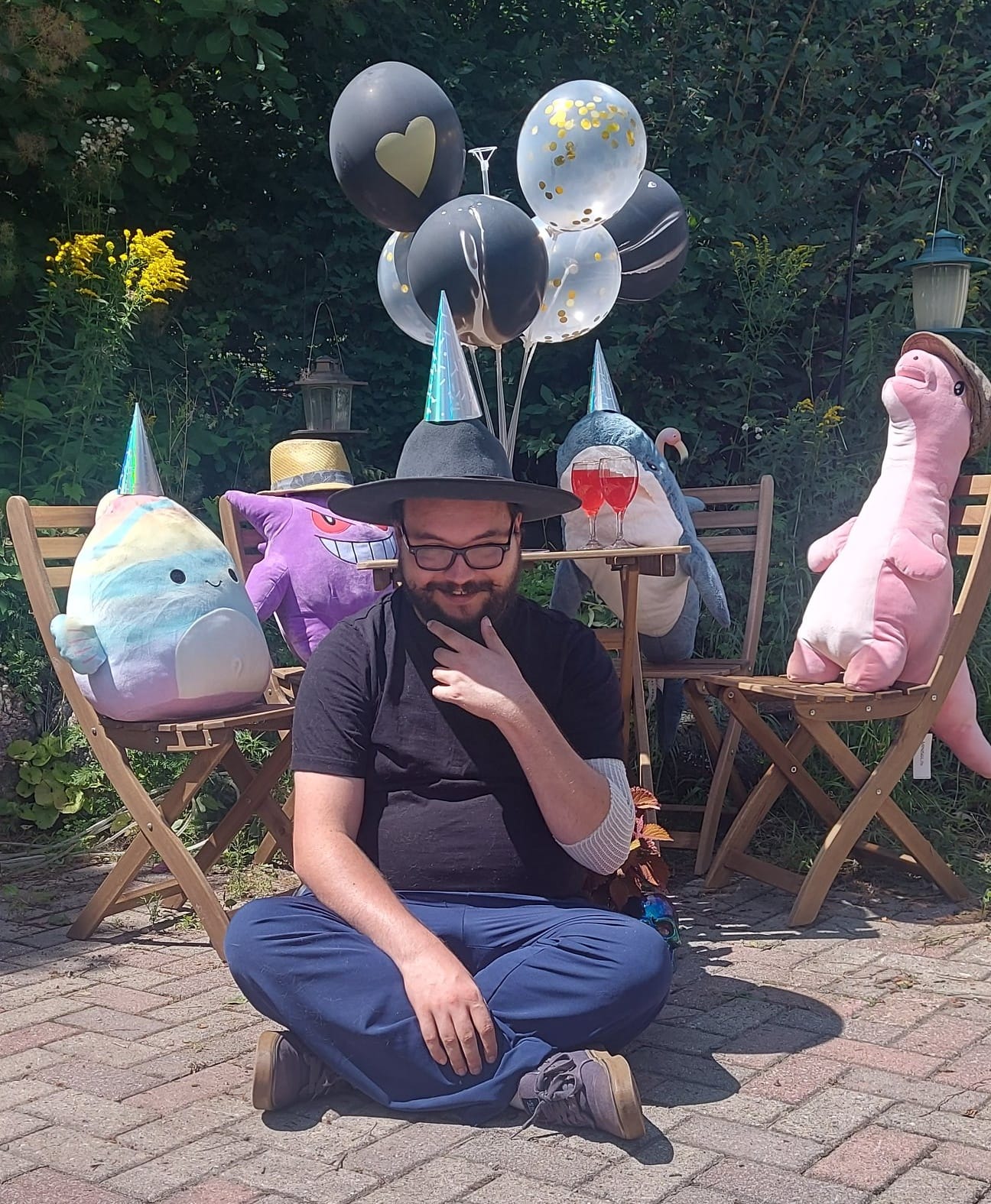 A man with facial hair and a felt sun hat sits on the ground in front of a patio set hosting a stuffed animal patio party, Photo 5